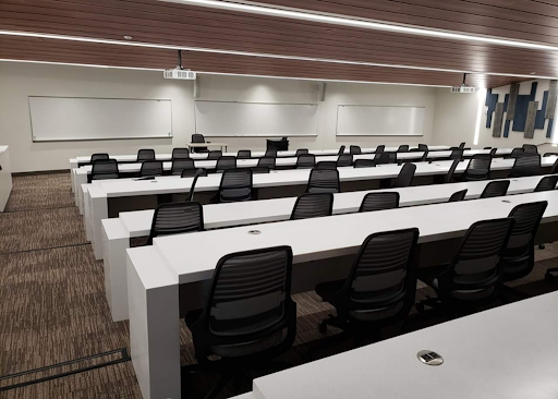 A lecture hall in Albertus Magnus Science Building with long white tables lined with swivel computer chairs, tiered with the highest level in the back, all facing the whiteboards at the low front of the room.