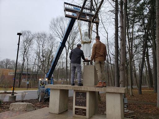 Two people stand on a raised platform guiding the statue of Our Lady of Fatima onto a pedestal. The statue is held by a crane.